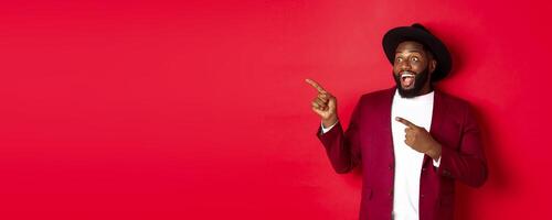 Winter holidays and shopping concept. Happy Black man pointing left and smiling, showing new year promo offer on red background photo