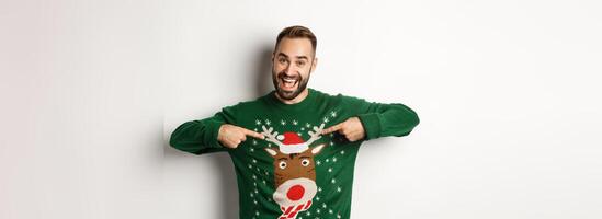 New year celebration and winter holidays concept. Cheerful man pointing at his funny Christmas sweater and smiling, white background photo