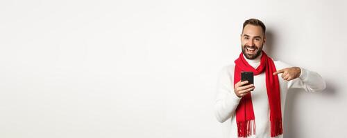 Satisfied bearded guy pointing finger at smartphone, checking out internet promo offer on mobile phone, standing over white background photo