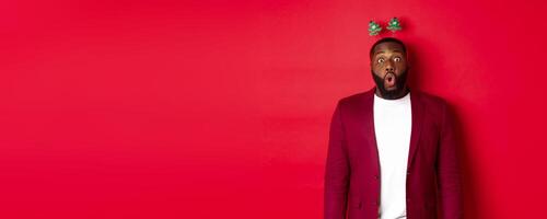 Merry Christmas. Happy african american man celebarting New Year, wearing party headband and making funny faces , standing over red background photo