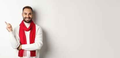 Christmas shopping and winter holidays concept. Young man with beard pointing finger left, showing new year advertisement, standing over white background in red scarf and sweater photo