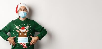 Concept of covid-19 and Christmas holidays. Funny guy put on face mask on his sweater deer, standing over white background photo