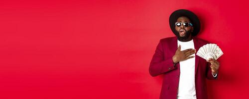 Shocked african american man holding hand on heart and gasping from excitement, showing huge amount of money, winning prize, standing over red background photo