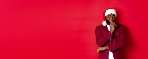 Christmas, party and holidays concept. Smiling african american man making new year plans, looking thoughtful at upper left corner, wearing santa hat, red background photo