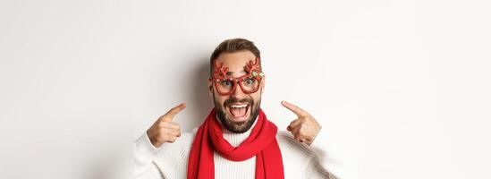 Christmas, New Year and celebration concept. Excited bearded man pointing at his party glasses, having fun and standing over white background photo