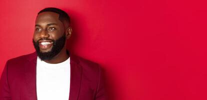 Close-up of handsome african american man with beard, looking left and smiling joyful, standing over red background photo
