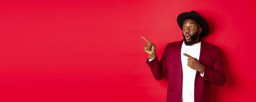 Winter holidays and shopping concept. Happy Black man pointing left and smiling, showing new year promo offer on red background photo