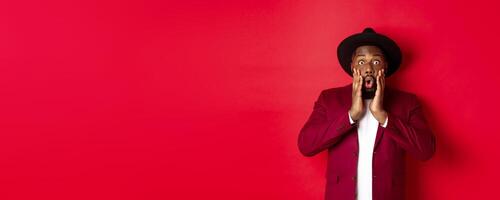Fashion and party concept. Impressed Black man in classy outfit staring with complete disbelief at camera, gasping and looking surprised, standing over red background photo