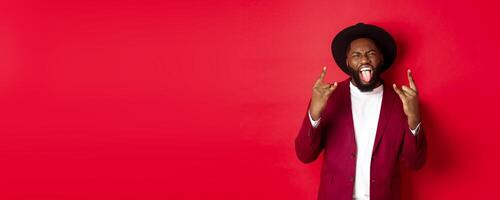Happy african american man having fun on party, showing tongue and rock-n-roll gesture, celebrating New Year, standing against red background photo