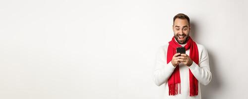 Man reading messaging on mobile phone and looking happy, standing in winter sweater and red scarf, white background photo