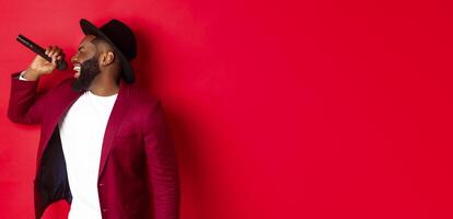 Passionate black male singer performing against red background, singing into microphone, wearing party outfit, standing over red background photo