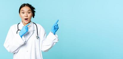 Shocked asian doctor, nurse in uniform, pointing right and gasping surprised, big news, standing over blue background photo