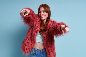 Beautiful young woman in fluffy coat pointing camera and smiling against blue background photo