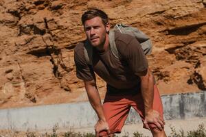Tired adventurer with backpack holding hands on knees while resting against a rocky mountain photo