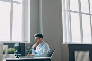 Confident young businessman using computer while sitting at his working place in office photo