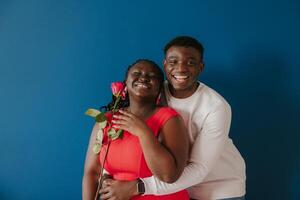 Happy young African man embracing his girlfriend holding red rose on blue background photo