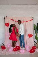 Smiling young African couple holding hands while standing on pink fabric background with heart shaped balloons photo