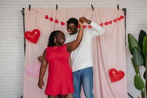 Smiling young African couple dancing on pink fabric background with heart shaped balloons photo