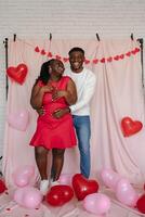 Full length of happy young African couple embracing on pink background with heart shaped balloons photo