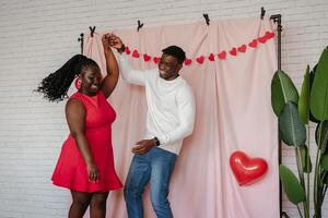 Joyful young African couple dancing on pink fabric background with heart shaped balloons photo