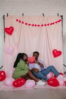 Romantic African couple covering faces with heart shaped Valentine card while sitting on pink fabric background photo