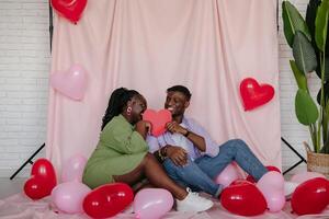 Smiling African couple covering faces with heart shaped Valentine card while sitting on pink fabric background photo