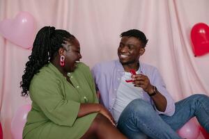 Happy young African man giving engagement ring to his girlfriend and asking to marry him on pink fabric background photo