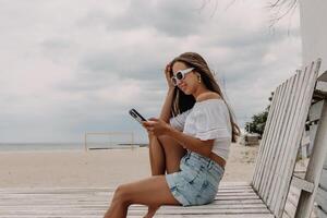 Happy young woman using smart phone while sitting on the wooden pallet seaside photo