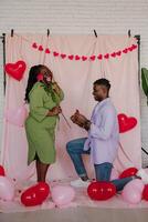 Young African man giving engagement ring to his surprised girlfriend while making proposal on pink background photo