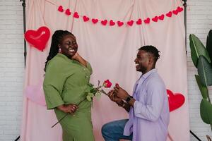 Handsome African man giving engagement ring to his surprised girlfriend while making proposal on pink background photo
