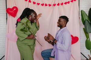 Young African man giving engagement ring to his surprised girlfriend while making proposal on pink background photo