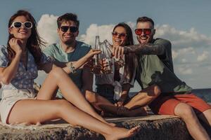 Group of happy young friends toasting with beer while spending carefree time seaside together photo