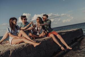Group of young friends toasting with beer and smiling while spending time seaside together photo