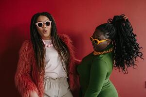 Two playful plus size African woman with braided hair having fun while standing on red background together photo