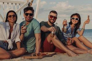 Four happy young friends looking at camera while enjoying pizza and beer on the beach photo