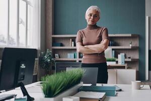 Senior businesswoman keeping arms crossed while standing near her working place in office photo
