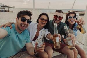 Group of cheerful young people making selfie while enjoying beer on the beach together photo