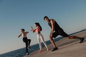 Three young people in sportswear doing stretching exercises before training seaside together photo