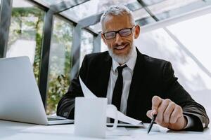 Mature businessman pointing paper with pen and smiling while analyzing graphs and charts in the office photo
