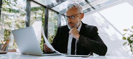 Confident mature businessman going through graphs and using technologies while working in office photo