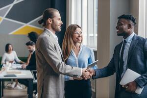 Two smiling young businessmen shaking hands in office while their colleagues working on background photo