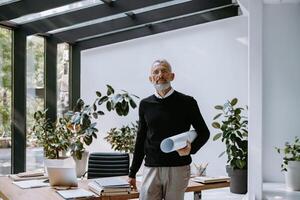 Confident mature architect carrying blueprint and looking at camera while standing in the office photo