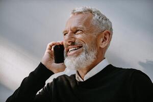 Portrait of happy senior man talking on mobile phone and smiling while standing against the wall photo