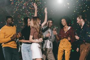Group of young joyful people enjoying champagne and throwing confetti while dancing in night club together photo