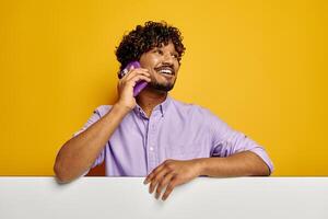 Happy Indian man leaning on the copy space and talking on phone against yellow background photo