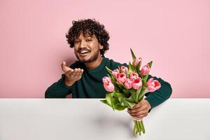 Handsome Indian man blowing a kiss while holding flower bouquet against pink background photo