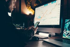 Close-up of confident young male stock trader using smart phone and computers while working late in office photo