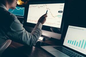 Close-up of male financial broker pointing computer monitor while analyzing stock market graph in night office photo