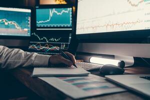 Close-up of male trader making notes and using computers while analyzing stock market data in night office photo