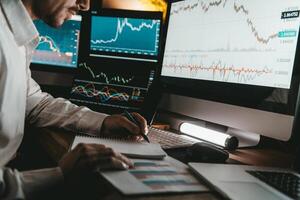 Close-up of male trader making notes and using computers while analyzing stock market data in night office photo